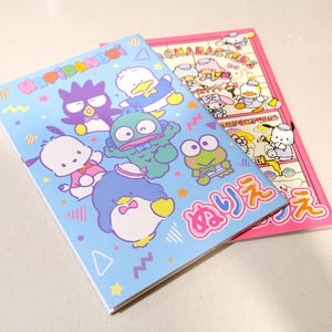 May include: A blue and pink notebook with a cartoon character design. The cover features a group of cartoon characters, including a penguin, a frog, a duck, and a monster. The notebook has the text "Happy Friends" and "みんな" on the cover.