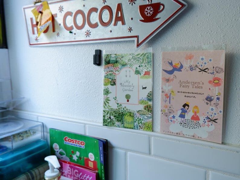 May include: Two framed illustrations on a white tiled wall. The illustration on the left is a colorful garden scene with the text "My Heavenly Garden". The illustration on the right is a pink and white scene with the text "Andersen's Fairy Tales" in a white banner.