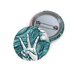 May include: A teal blue and white button featuring a woman's face with her hand making a peace sign. The woman's hair is flowing behind her.