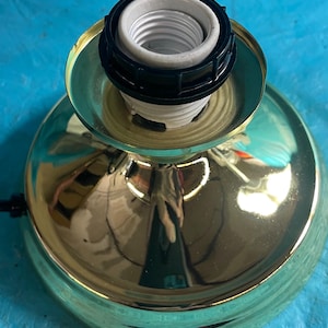 Puede incluir: Una base de lámpara dorada con un casquillo de bombilla negro y blanco. La base tiene una superficie brillante y reflectante y una forma redondeada. El casquillo está diseñado para alojar una bombilla estándar.