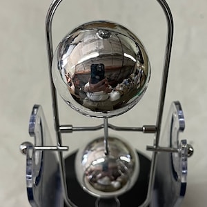 Könnte beinhalten: Ein Newtonsches Wiege-Schreibtischspielzeug mit schwarzem Sockel, transparenten Kunststoffstützen und glänzenden silbernen Metallkugeln. Die Kugeln sind aufgehängt und reflektieren die Umgebung. Ein klassisches Geschenk.