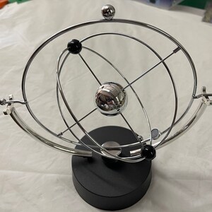Puede incluir: Una cuna de Newton cromada con una base negra. La escultura cinética presenta una gran esfera central, esferas orbitales más pequeñas y una serie de arcos metálicos interconectados. El diseño es una versión moderna de un juguete de escritorio clásico.