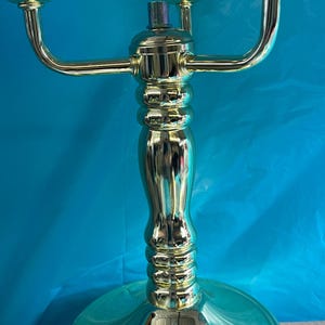 Può includere: Un candelabro dorato con tre portalampada. La base è rotonda e lucida, e lo stelo ha un design curvo decorativo. Il candelabro è in metallo e ha una finitura lucida. Lo sfondo è di colore turchese.