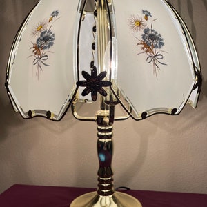 Può includere: Lampada da tavolo vintage con base e stelo in ottone, sormontata da un paralume a motivi floreali. Il paralume presenta uno sfondo bianco con illustrazioni di margherite e fiori blu. La lampada ha un accento floreale nero decorativo.