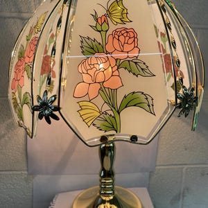 Puede incluir: Una lámpara de mesa decorativa con base y tallo dorados. La pantalla es octogonal, con un fondo crema y un diseño floral con rosas rosas, hojas verdes y una mariposa amarilla. La pantalla está adornada con metal dorado.