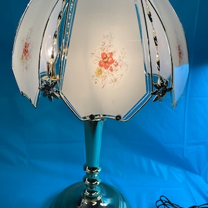 Pode incluir: Uma lâmpada de mesa vintage com um design floral no abajur. A lâmpada apresenta uma base e detalhes dourados, uma haste azul clara e um abajur de vidro fosco com flores vermelhas e amarelas. A lâmpada está ligada e acesa.