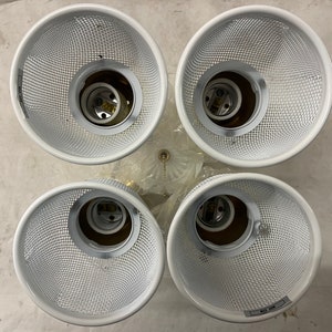 May include: Overhead view of a four-light fixture with white mesh shades. The fixture has a brass-colored base and is designed to hold four light bulbs. The shades are cylindrical and have a grid-like pattern.