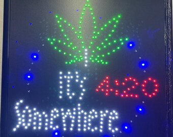 Puede incluir: Letrero LED negro con una hoja de cannabis verde, el texto "It's 4:20 Somewhere" en blanco y rojo. El letrero tiene un marco negro y luces LED azules alrededor del borde.