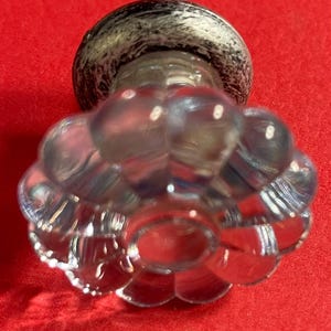 May include: A clear glass bottle stopper with a flower-like design. The stopper has a rounded top with a black and white marbled pattern. The base of the stopper has a series of clear, rounded shapes, creating a floral appearance.