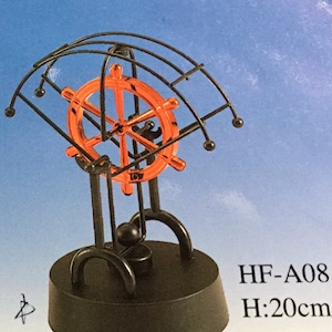 Puede incluir: Escultura cinética decorativa con un volante de barco naranja brillante dentro de un marco de metal negro. La base es negra y la altura es de 20 cm. La escultura tiene un diseño equilibrado con una pequeña bola negra suspendida.