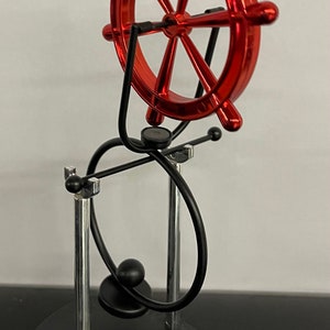 Puede incluir: Escultura de timón rojo con base negra y plateada. El timón es de metal rojo y tiene un acabado brillante. La escultura tiene un diseño único con soportes de metal negro curvos y una base negra.