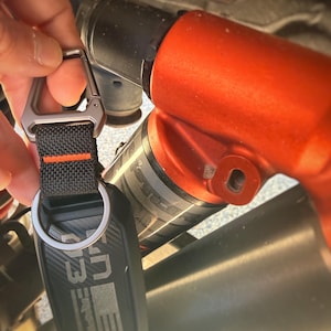 Puede incluir: Un llavero negro y gris con un dise&ntilde;o de fibra de carbono, sujeto a una correa negra con una raya naranja y un mosquet&oacute;n plateado. El llavero tiene el texto "UTV" y "3".