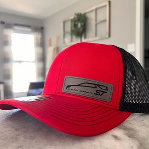 May include: Red and black trucker hat with a mesh back. The front of the hat features a rectangular patch with the text "EXPLORER ST" and a car silhouette. The hat has a curved brim.