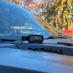 Puede incluir: Un llavero negro y naranja con el texto "CN 03" y un dise&ntilde;o de bandera estadounidense descansa sobre el cap&oacute; de una camioneta Raptor gris. La palabra "RAPTOR" se muestra en letras negras. El fondo muestra &aacute;rboles y un cielo azul claro.
