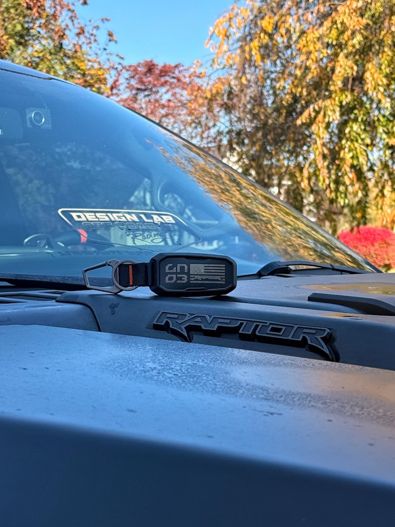 Puede incluir: Un llavero negro y naranja con el texto "CN 03" y un dise&ntilde;o de bandera estadounidense descansa sobre el cap&oacute; de una camioneta Raptor gris. La palabra "RAPTOR" se muestra en letras negras. El fondo muestra &aacute;rboles y un cielo azul claro.