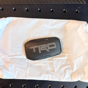 May include: A rectangular automotive emblem featuring a black carbon fiber pattern with the letters TRD embossed in the center. The emblem has a black border and is resting on a piece of white crinkled paper, showcasing a textured finish.