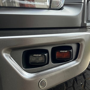 May include: Close-up of a gray truck's rear bumper with two black and red LED lights. The lights are labeled "RIGID" and are mounted in the bumper.