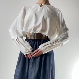 Op de afbeelding: Een witte blouse met lange mouwen en een sleutelgat-halslijn, gecombineerd met een marineblauwe rok. De blouse heeft geplooide details op de mouwen en wordt in de taille vastgehouden met een brede gouden riem. Een gouden ketting is ook zichtbaar.
