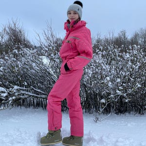 Puede incluir: Un mono de esquí rosa brillante con capucha a juego y gorro gris con pompón negro. El mono tiene cremallera frontal, bolsillos y ribetes grises. La persona lleva botas de nieve verde oliva. El fondo es un paisaje nevado.