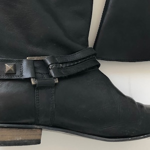 May include: A black leather boot featuring a low heel with a light brown wood-tone finish. The boot includes a decorative ankle strap adorned with metallic pyramid-shaped studs and a rectangular buckle, showcasing a smooth texture and rounded toe.