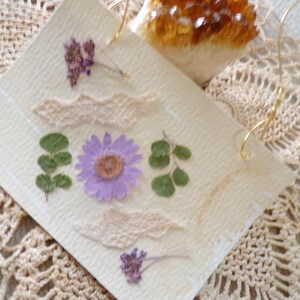 May include: A white card with pressed flowers and leaves. The flowers are purple and white, and the leaves are green. The card is on a white crocheted surface.