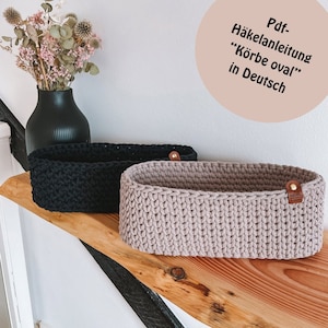 Könnte beinhalten: Zwei gehäkelte ovale Körbe, einer schwarz und einer hellbraun, auf einem Holzregal. Die Körbe haben Ledergriffe und sind mit einem strukturierten Stich gearbeitet. Der Text "Pdf-Häkelanleitung "Körbe oval" in Deutsch" ist in der oberen rechten Ecke des Bildes sichtbar.