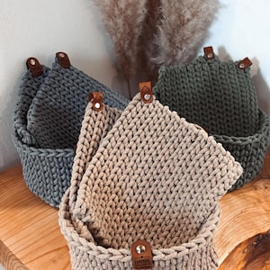 Könnte beinhalten: Drei gehäkelte Aufbewahrungskörbe mit Ledergriffen. Die Körbe sind in den Farben Grau, Grün und Beige. Der beige Korb hat ein "Handmade"-Etikett.