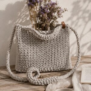 May include: A light gray hand-knitted tote bag with a thick cord texture and a single brown leather tab detail. The bag features a long shoulder strap and is filled with a bouquet of dried purple and white statice flowers, creating a soft aesthetic.