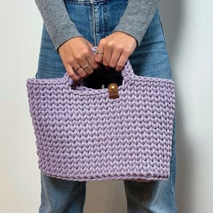 May include: A light purple crochet handbag featuring a thick, textured knit pattern and two integrated handles. A small, rectangular brown leather-like tag with a gold-colored metal rivet is attached to the top center of the bag.