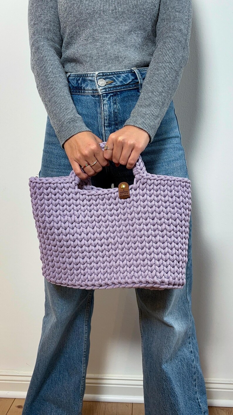 May include: A light purple crochet handbag featuring a thick, textured knit pattern and two integrated handles. A small, rectangular brown leather-like tag with a gold-colored metal rivet is attached to the top center of the bag.