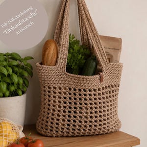 Könnte beinhalten: Eine beige gehäkelte Einkaufstasche gefüllt mit Lebensmitteln, darunter ein Brotlaib, grünes Gemüse und eine Packung Kichererbsen. Die Tasche hat eine rechteckige Form mit einem gewebten Muster und stabilen Griffen. Der Text auf dem Bild lautet "Pdf Häkelanleitung "Einkaufstasche" in Deutsch."