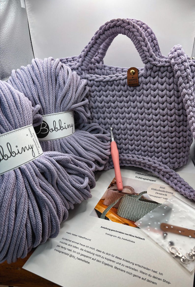 May include: A lavender crocheted handbag with a brown leather-like tag, two skeins of 5mm Bobbiny premium cord, a pink crochet hook, a printed instruction sheet titled H&auml;kelanleitung Fr&uuml;hlingsshopper, and a small plastic bag with metal hardware.