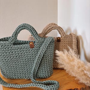 May include: Two crocheted handbags, one in sage green and one in beige, featuring a thick cord texture, sturdy handles, and a small brown leather-like tag with a gold rivet on the front center of each bag, accompanied by a long shoulder strap.
