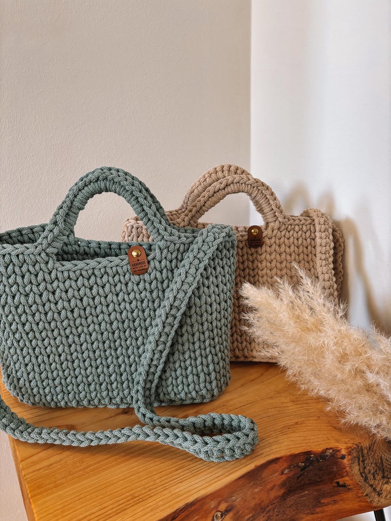 May include: Two crocheted handbags, one in sage green and one in beige, featuring a thick cord texture, sturdy handles, and a small brown leather-like tag with a gold rivet on the front center of each bag, accompanied by a long shoulder strap.