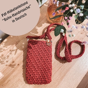 May include: A red crocheted phone bag with a brown leather tag that says "Handmade with Love". The bag has a long strap and is sitting on a wooden surface.