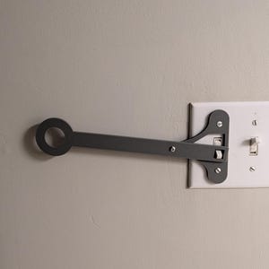 May include: A dark gray metal light switch extender with a circular handle. The extender is attached to a white light switch plate on a light gray wall. The switch is in the on position.