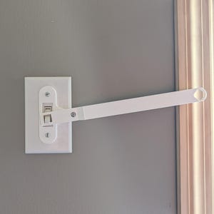 May include: A white light switch extender installed on a white light switch plate. The extender has a long, straight arm with a loop at the end, designed to make it easier to turn the light switch on and off. The light switch plate is mounted on a gray wall.