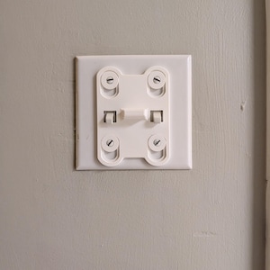 May include: A white plastic light switch guard mounted over a standard rectangular wall plate. The guard features a central horizontal bar that covers the toggle switch, secured by four circular screw-in fasteners at each corner of the frame.