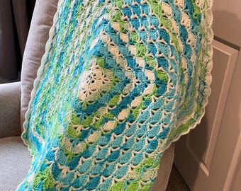 May include: A handmade crochet blanket in shades of blue, green, and white, draped over a gray chair. The blanket features a shell stitch pattern, creating a textured surface. The blanket is approximately 40 inches wide and 40 inches long.