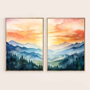 May include: Two framed watercolor landscape paintings. Each features a mountain range with a sunset sky in shades of orange and pink. The foreground shows dark green trees and blue-toned mountains.
