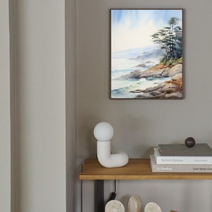 May include: A framed watercolor painting of a coastal landscape with trees, rocks, and ocean waves. The painting is displayed on a gray wall above a wooden shelf with a white lamp, books, and decorative objects. The scene evokes a sense of tranquility.