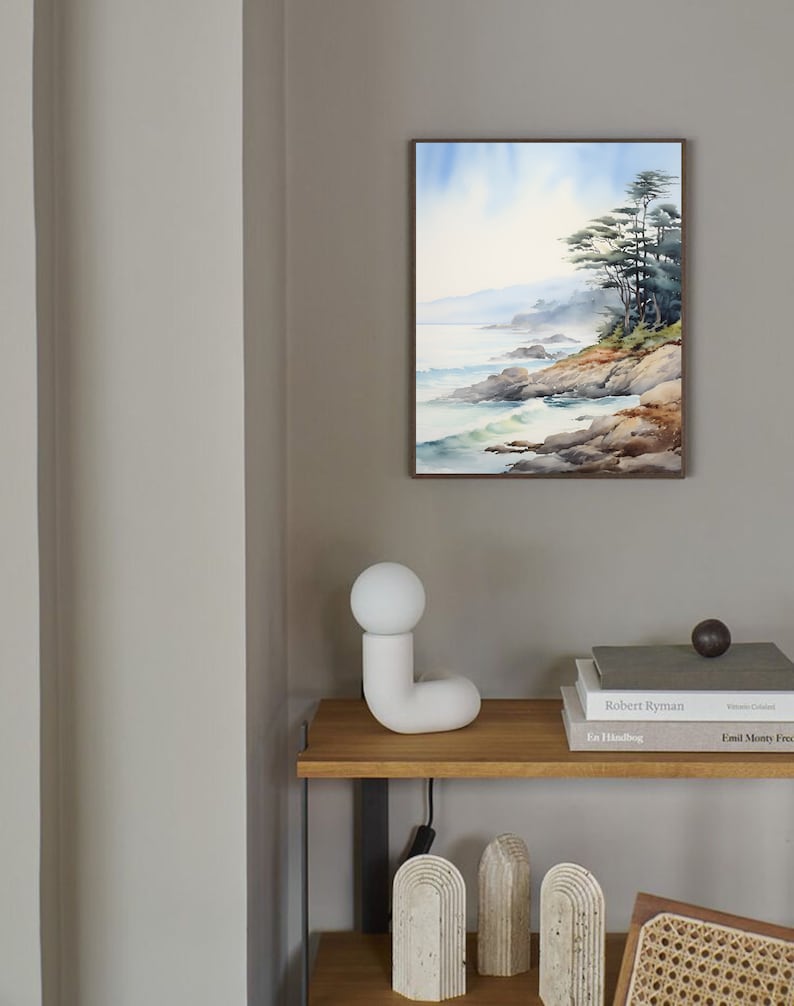 May include: A framed watercolor painting of a coastal landscape with trees, rocks, and ocean waves. The painting is displayed on a gray wall above a wooden shelf with a white lamp, books, and decorative objects. The scene evokes a sense of tranquility.