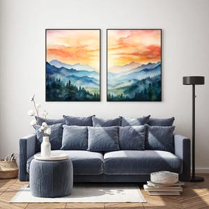 May include: Two framed watercolor landscape paintings depict mountain ranges and forests under a vibrant orange and yellow sky. The artwork hangs above a blue sofa with matching pillows, a round ottoman, and a floor lamp.