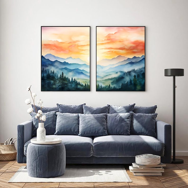 May include: Two framed watercolor landscape paintings depict mountain ranges and forests under a vibrant orange and yellow sky. The artwork hangs above a blue sofa with matching pillows, a round ottoman, and a floor lamp.
