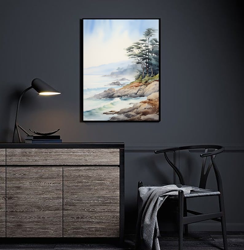 May include: Watercolor painting of a coastal landscape with a black frame. The artwork depicts a rocky shoreline, ocean waves, and evergreen trees under a soft blue sky. The painting is displayed on a dark gray wall above a wooden cabinet.