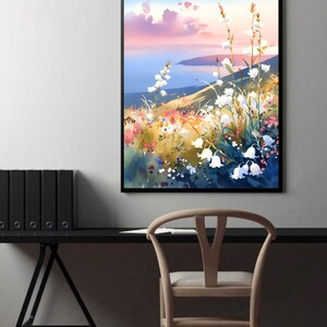 Pode incluir: Uma pintura de paisagem em aquarela emoldurada com uma colina com flores brancas, flores silvestres coloridas e um corpo de &aacute;gua sob um c&eacute;u azul e rosa. A obra de arte &eacute; exibida em uma parede branca acima de uma mesa e cadeira pretas.