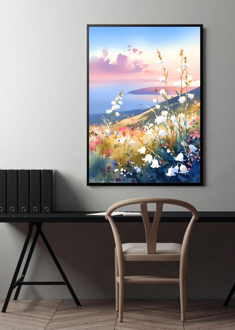 Pode incluir: Uma pintura de paisagem em aquarela emoldurada com uma colina com flores brancas, flores silvestres coloridas e um corpo de &aacute;gua sob um c&eacute;u azul e rosa. A obra de arte &eacute; exibida em uma parede branca acima de uma mesa e cadeira pretas.