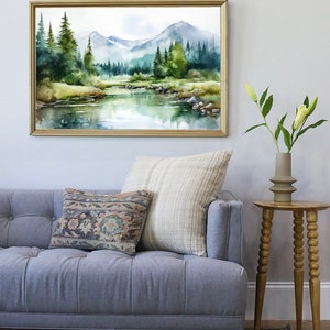 May include: A watercolor painting of a mountain landscape with a river flowing through it. The painting is framed in a gold frame and is hanging on a white wall. There is a gray sofa with pillows in front of the painting.