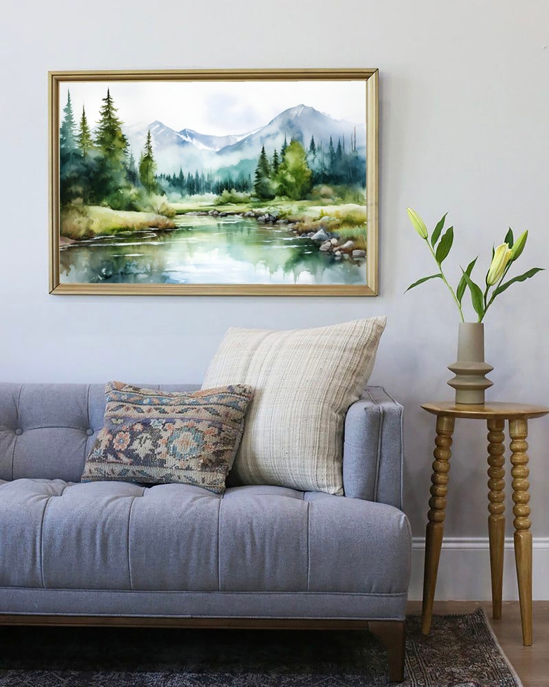 May include: A watercolor painting of a mountain landscape with a river flowing through it. The painting is framed in a gold frame and is hanging on a white wall. There is a gray sofa with pillows in front of the painting.