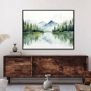 May include: Watercolor painting of a lake reflecting a mountain range and a forest with green trees. The painting is framed in black and hanging on a white wall above a wooden cabinet.